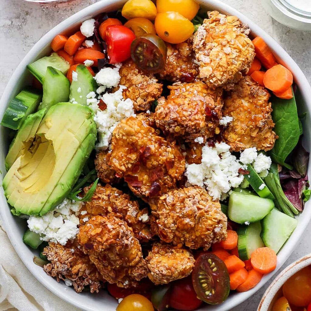 A bowl containing fried chicken, avocado slices, cherry tomatoes, sliced cucumbers, carrots, and leafy greens, topped with crumbled cheese.