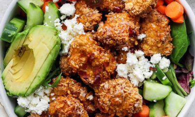 A bowl containing fried chicken, avocado slices, cherry tomatoes, sliced cucumbers, carrots, and leafy greens, topped with crumbled cheese.