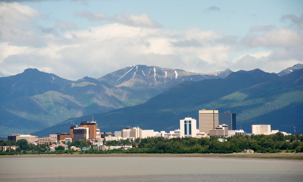 A panoramic view of Anchorage, Alaska, showcasing the city skyline with tall buildings against the backdrop of the Chugach Mountains and a cloudy sky.