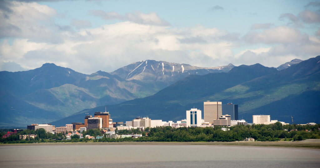 A panoramic view of Anchorage, Alaska, showcasing the city skyline with tall buildings against the backdrop of the Chugach Mountains and a cloudy sky.