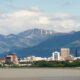 A panoramic view of Anchorage, Alaska, showcasing the city skyline with tall buildings against the backdrop of the Chugach Mountains and a cloudy sky.