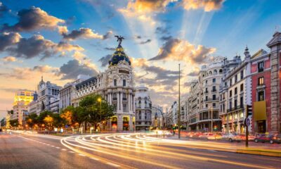 A bustling street in Madrid at dusk, with long exposure capturing streaks of light from moving traffic. Historic buildings with intricate facades line the avenue, while the sky above shows a dynamic pattern of clouds illuminated by crepuscular rays.