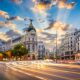 A bustling street in Madrid at dusk, with long exposure capturing streaks of light from moving traffic. Historic buildings with intricate facades line the avenue, while the sky above shows a dynamic pattern of clouds illuminated by crepuscular rays.