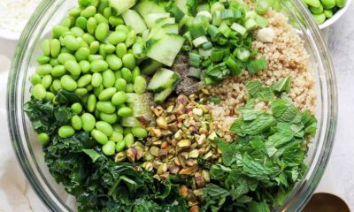 A bowl of salad containing quinoa, kale, chopped cucumbers, green onions, shelled edamame, chopped nuts, and fresh herbs, with a pair of golden spoons on the.