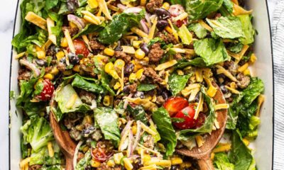 A colorful taco salad with lettuce, tomatoes, ground beef, cheese, and black beans served in a rectangular dish with wooden serving utensils.