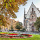 A historic church with a large, ornate entrance sits behind a circular garden with colorful flowers and vibrant autumn foliage.