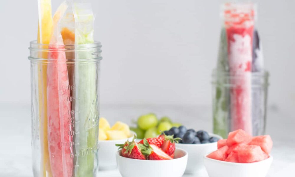 Two glass jars of homemade fruit popsicles sit on a counter alongside small white dishes of fruit.