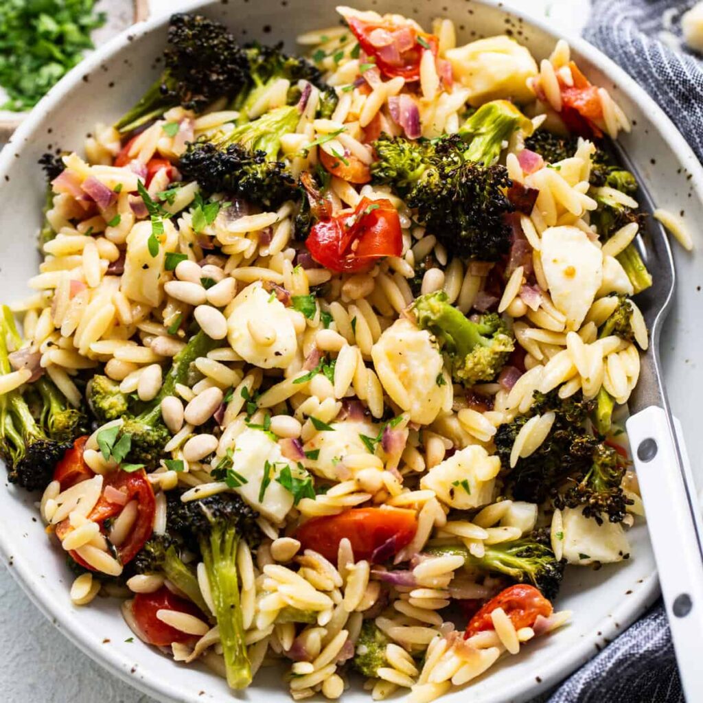 A bowl of orzo pasta salad with roasted broccoli, cherry tomatoes, and pine nuts, garnished with herbs.