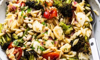 A bowl of orzo pasta salad with roasted broccoli, cherry tomatoes, and pine nuts, garnished with herbs.