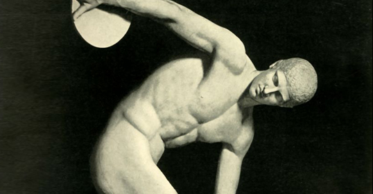 Black-and-white photo of a discus thrower sculpted in ancient Greek style, depicting a muscular male figure in mid-throw, symbolizing principles of physical fitness.