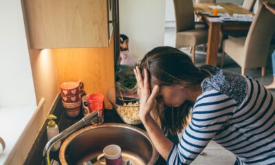 Stressed mum at home. She has her head in her hands at a messy kitchen sink and her children are running round in the background.