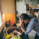 Stressed mum at home. She has her head in her hands at a messy kitchen sink and her children are running round in the background.