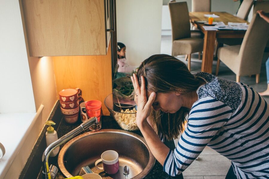 Stressed mum at home. She has her head in her hands at a messy kitchen sink and her children are running round in the background.