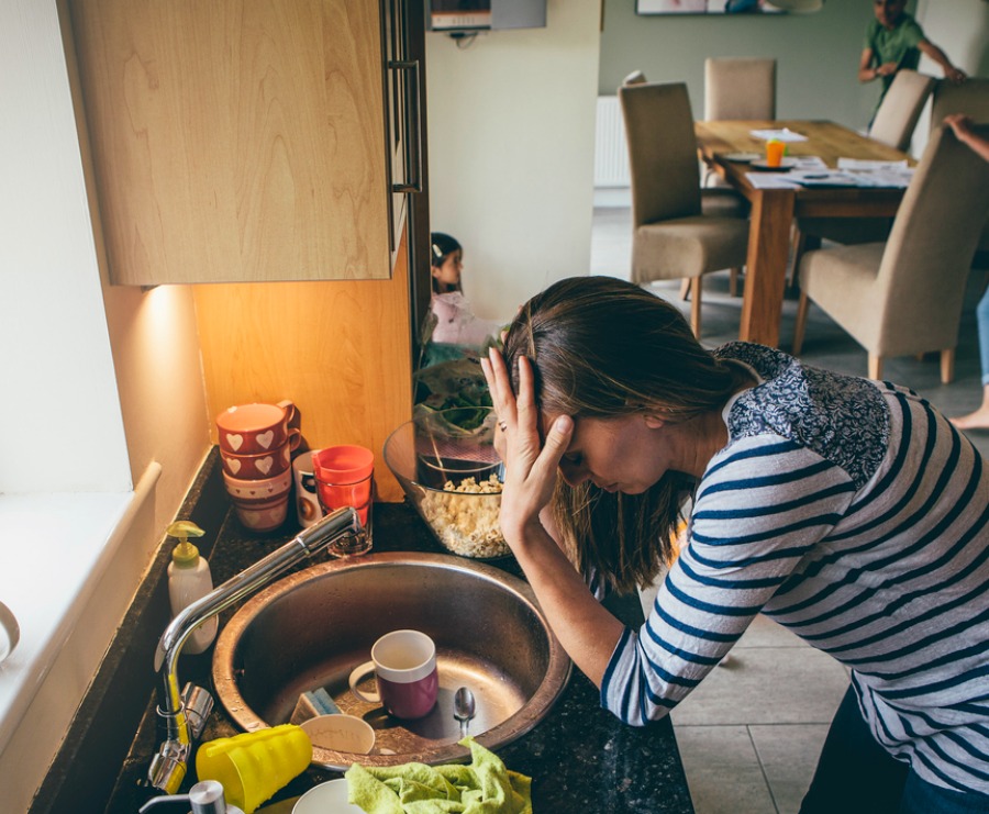 Stressed mum at home. She has her head in her hands at a messy kitchen sink and her children are running round in the background.