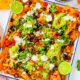 Loaded veggie nachos piled with veggies, jalapenos, guacamole, sour cream, cheese, olives served on a white platter.