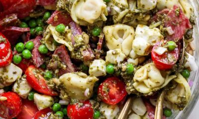 A close-up of a pasta salad with tortellini, cherry tomatoes, peas, mozzarella balls, salami, and green pesto sauce, served in a glass bowl with metal utensils.