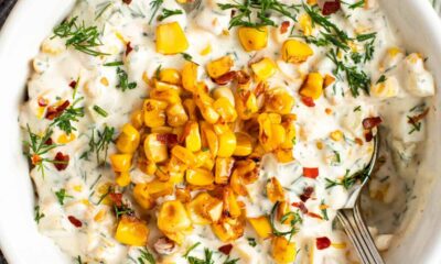 A bowl of creamy dip topped with roasted corn, chopped herbs, and red pepper flakes, with a spoon on the side.