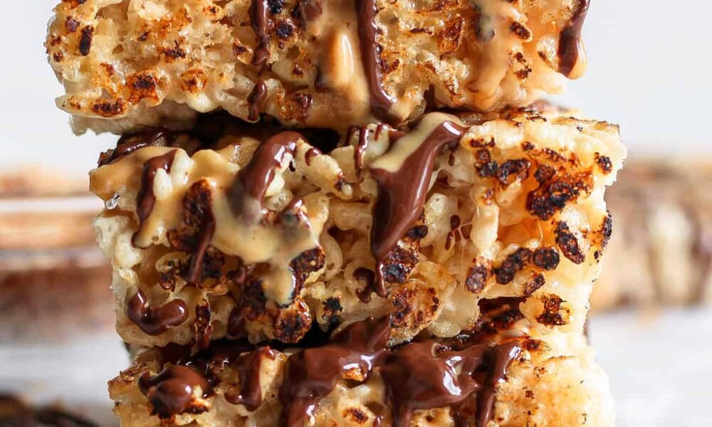 Stacked toasted rice krispie treats drizzled with chocolate and caramel sauces on a white surface.