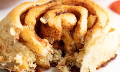 A close-up view of a partially eaten cinnamon roll on a white plate, showing its swirled layers and a bite taken out, with blurred background elements.