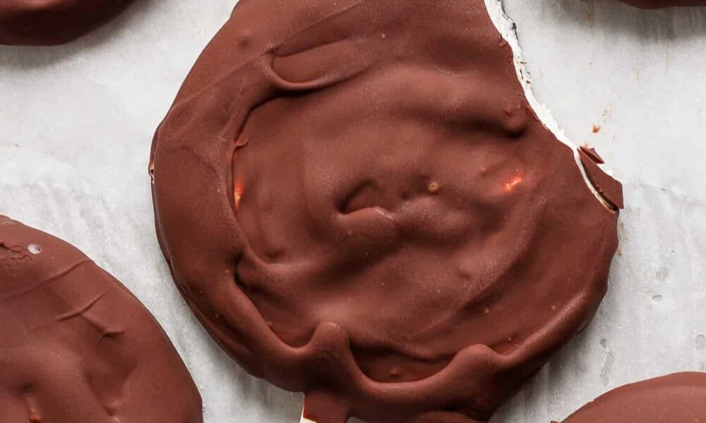 Close-up of a chocolate-covered round ice cream treat on a stick with a bite taken out of the edge, resting on parchment paper along with other similar treats.