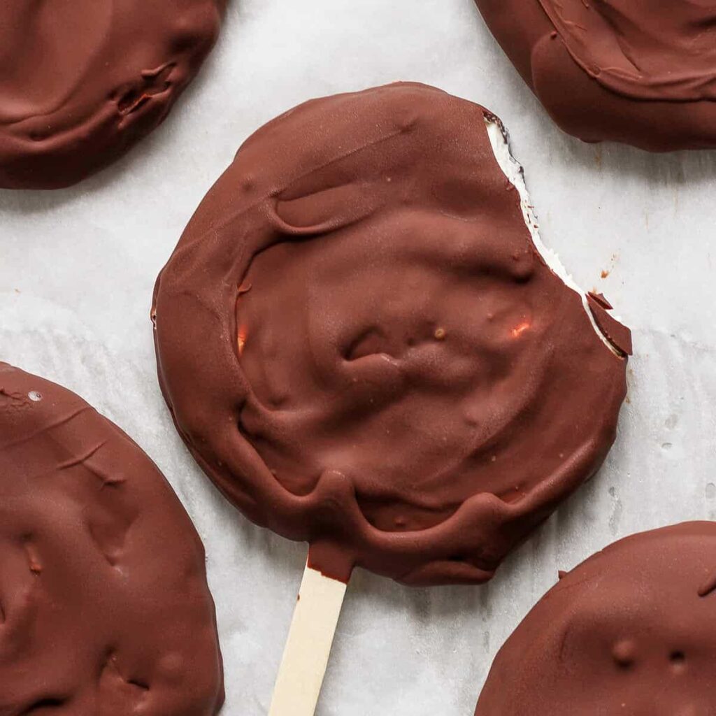 Close-up of a chocolate-covered round ice cream treat on a stick with a bite taken out of the edge, resting on parchment paper along with other similar treats.