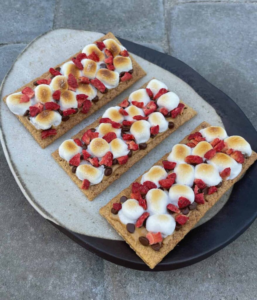 Three open-faced peanut butter and jelly s'mores dessert crackers on white ceramic serving platter.