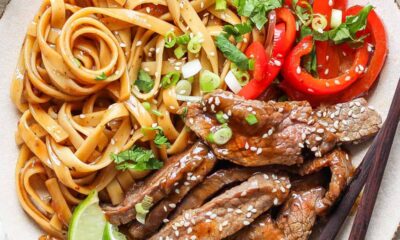 A plated dish featuring seasoned noodles, slices of red bell pepper, sesame-seeded beef strips, garnished with green onions and lime wedges, alongside a pair of chopsticks.