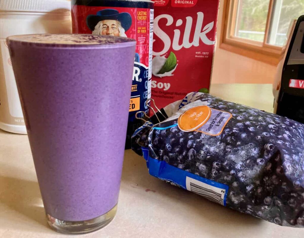 Tall full glass of blueberry Oatmeal Protein Shake surrounded by ingredients and blender.