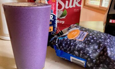 Tall full glass of blueberry Oatmeal Protein Shake surrounded by ingredients and blender.