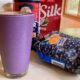 Tall full glass of blueberry Oatmeal Protein Shake surrounded by ingredients and blender.