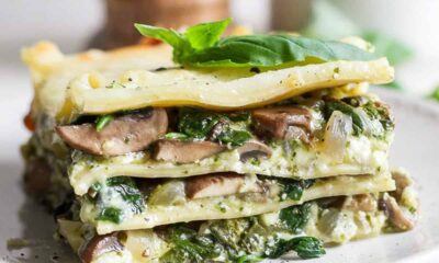 A slice of vegetable lasagna with visible layers of spinach, mushrooms, and cheese on a white plate, garnished with fresh basil leaves.