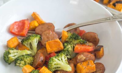 A white bowl of Sheet Pan Chicken Sausage & Veggies sits on a countertop with a brown linen napkin and a glass of water.