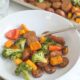 A white bowl of Sheet Pan Chicken Sausage & Veggies sits on a countertop with a brown linen napkin and a glass of water.