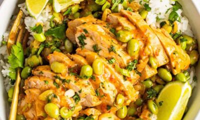 A bowl of skillet-cooked coconut chicken served over white rice, garnished with edamame beans, chopped herbs, and lime wedges.