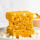 Three slices of golden yellow pumpkin blondies stacked on a plate, surrounded by white chocolate chips.