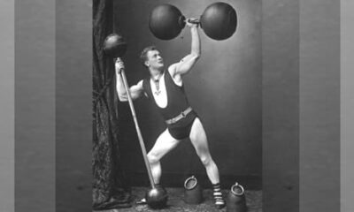 A man performs a bicep curl with a dumbbell on a beach while wearing shorts and a weightlifting belt, showcasing his dedication to fitness. Several people are in the background.