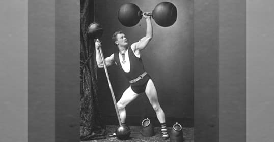 A man performs a bicep curl with a dumbbell on a beach while wearing shorts and a weightlifting belt, showcasing his dedication to fitness. Several people are in the background.